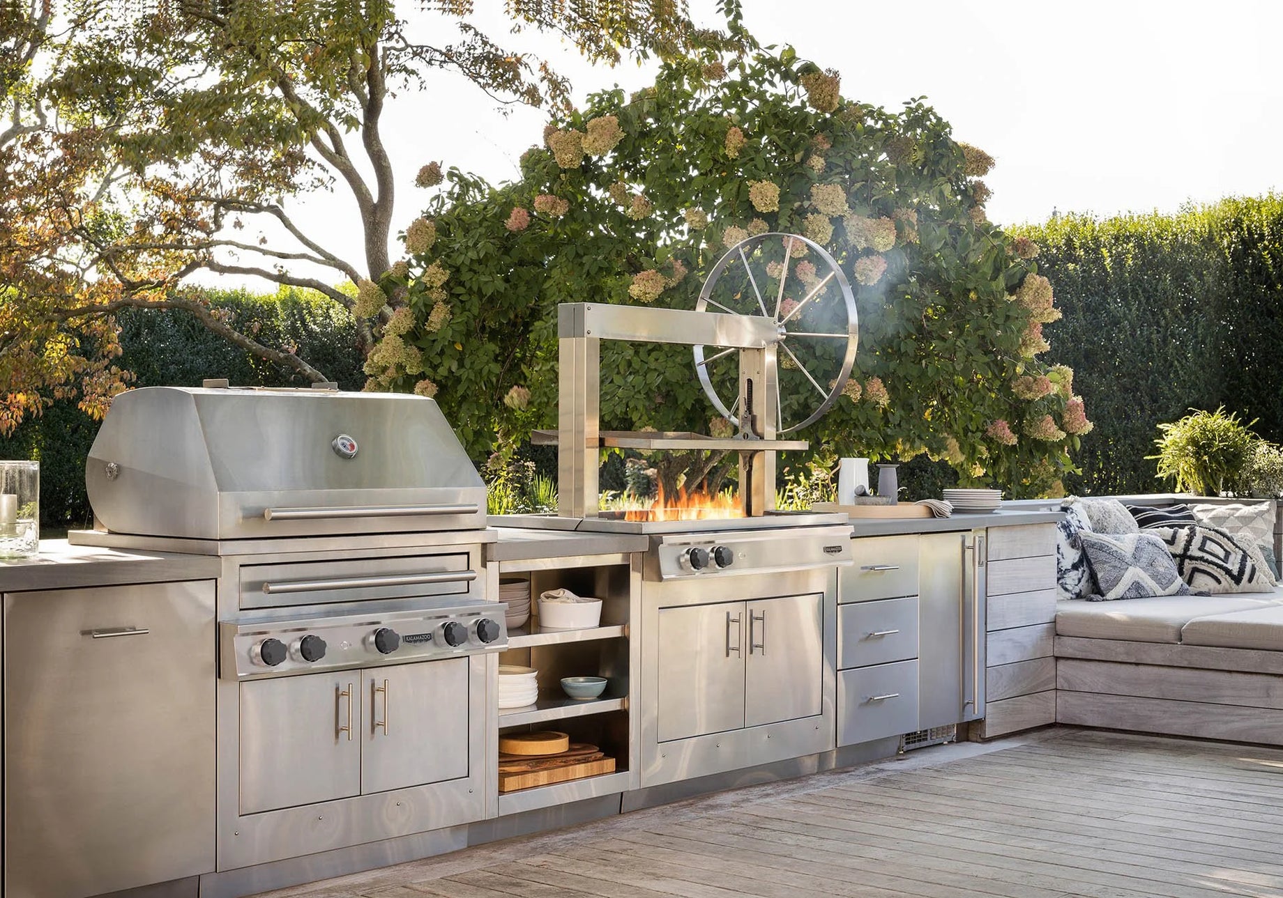 Outdoor kitchen with stainless steel appliances and a fire pit in a garden setting.